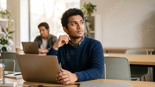 Thoughtful young Indian man working on laptop in modern office or coworking space. Pensive male student studying and looking away. Career and education concept.