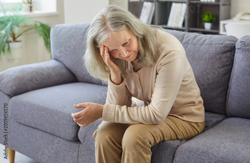 Sad elderly woman suffers from headache or migraine at home, sitting on sofa, facing health problem and pain, reflecting sick discomfort in a escape zone filled with senior care and distress.