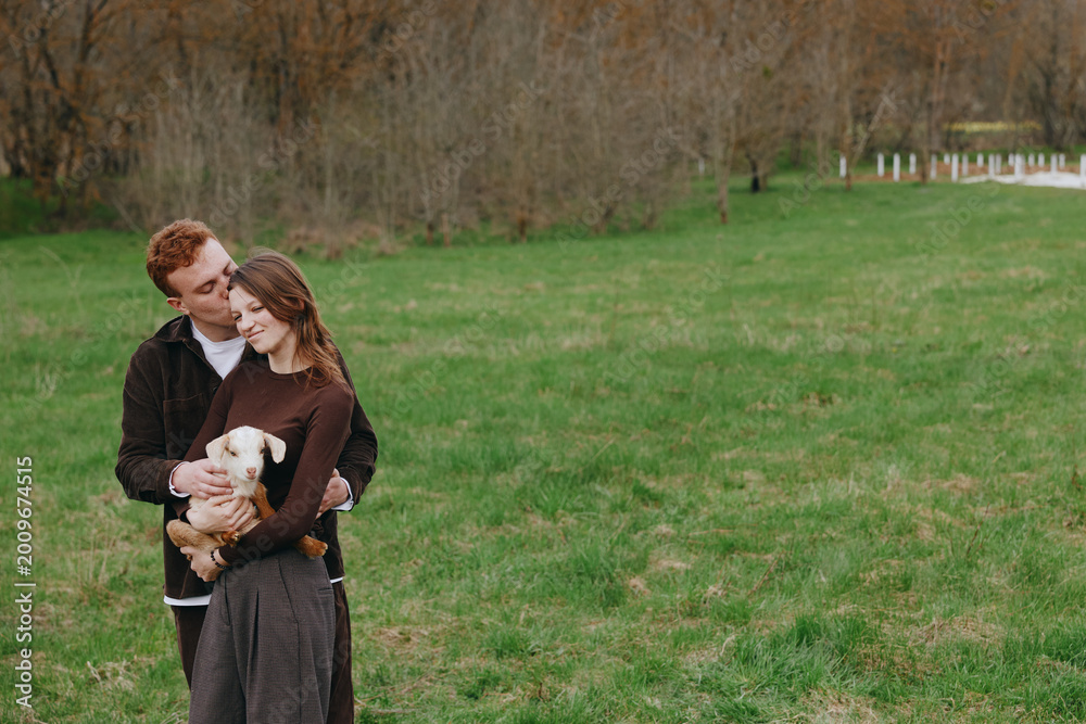 Naklejka premium A couple smiles in a sunlit park, one embracing a small dog. The scene captures warmth, companionship, and everyday romance in a bright, open green field.