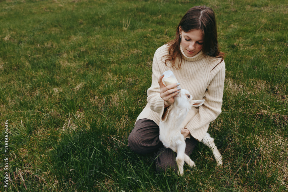 Fototapeta premium A woman sits on grass in a quiet field, bottle feeding a white goat kid, sharing a gentle intimate moment with a small animal, capturing a warm lifestyle scene amid nature and open space