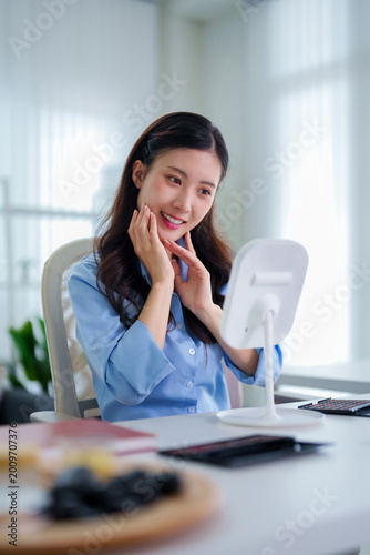 young Asian women admiring face using beauty mirror skincare routine glowing skin checking complexion sitting office desk morning natural light wellness