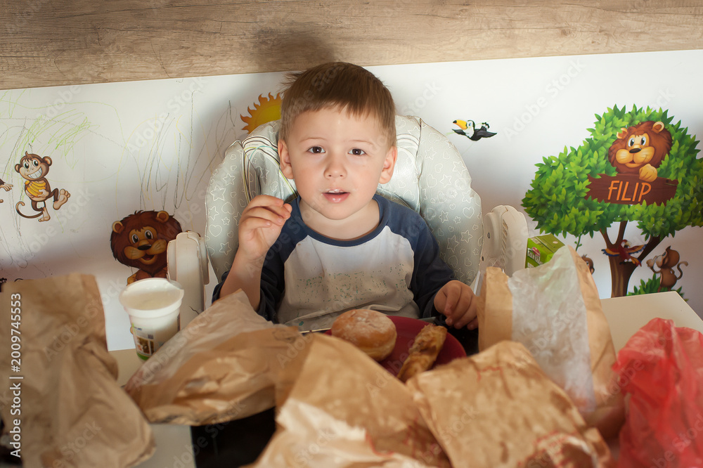 Fototapeta premium A child sits at a table enjoying breakfast. Food wraps and bags are scattered around. The room has animal wall art. The child looks up while holding a piece of food
