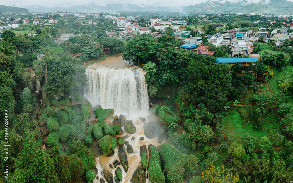 Fototapeta premium An aerial view of the stunning Thac Voi Waterfall in Da Lat, Vietnam, showcasing its powerful cascade amidst lush green surroundings and a nearby town. ,