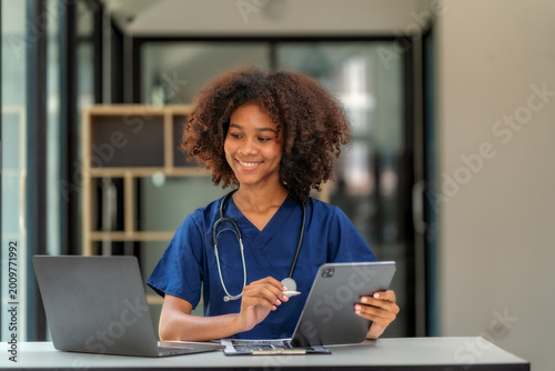 Smiling African American female doctor using digital tablet and laptop at clinic desk.
