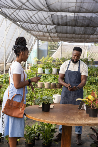 African American vendor and customer paying using phones at greenhouse table with tablet, plants