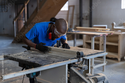 Wooden plank sliding through table saw in workshop with sawdust on concrete floor near high windows