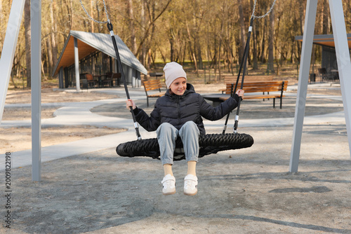 A child enjoys swinging on a tire swing in a public park during daytime. Nearby, there are trees and places for people to sit and relax