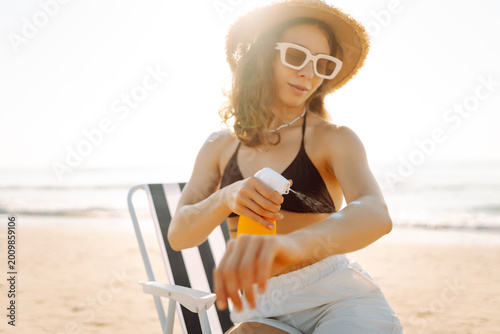 Woman in hat sits on sun lounger by water, applying sunscreen. Young woman on vacation applies sunscreen and enjoys sunset on beach. Concepts of protection, care, and enjoyment.