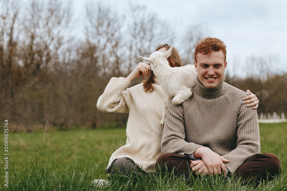 Fototapeta premium A couple enjoys an outdoor moment with a playful goat kid, sharing affection and laughter, sitting on grass in a park, showcasing a casual lifestyle and warm bond