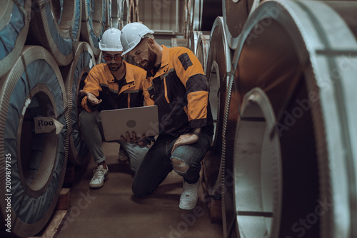 Bild auf Leinwand Diligent Surveyor in Steel Mill: A focused engineer, clad in protective gear, meticulously reviews data on his laptop amidst the massive rolls of steel, symbolizing precision and industrial expertise