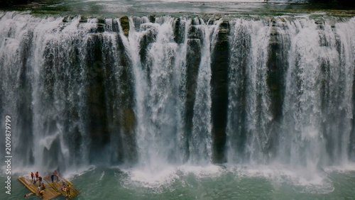 Majestic waterfall cascading into a serene turquoise pool, a powerful natural wonder. Lush green environment with a small raft of people, an adventurous outdoor experience.
