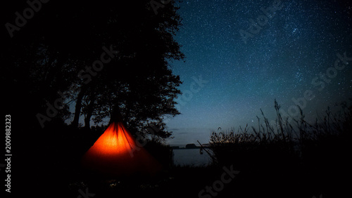 Starry night sky over a tranquil lake, an illuminated orange tent glowing under the vast dark cosmos, serene outdoor camping adventure, peaceful wilderness escape.