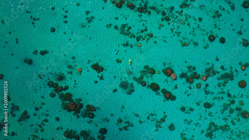 Aerial view of a lone kayaker paddling across vibrant turquoise ocean water, surrounded by scattered coral reefs and underwater rock formations, a serene tropical seascape for travel and nature