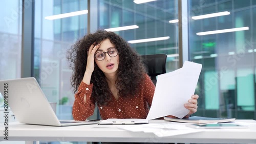 Frustrated businesswoman unhappy with financial results sitting at workplace in business office. Upset puzzled female financier unhappily reading document with bad indicators looking at laptop screen