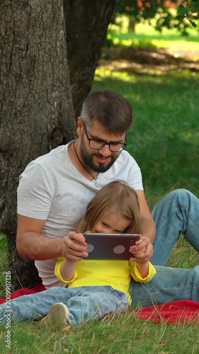 Happy family father and little girl sit in summer park with tablet. Computer games surfing internet social media. Picnic outdoors. Vertical shot