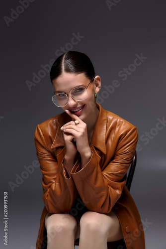 Stylish young woman in leather coat and glasses poses elegantly in studio setting