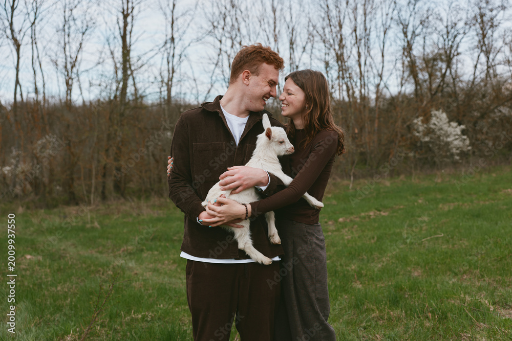 Fototapeta premium Cheerful couple outdoors, embracing a goat kid, smiling together in a sunlit field, calm countryside mood, natural colors, relaxed posture, tender connection, casual fashion