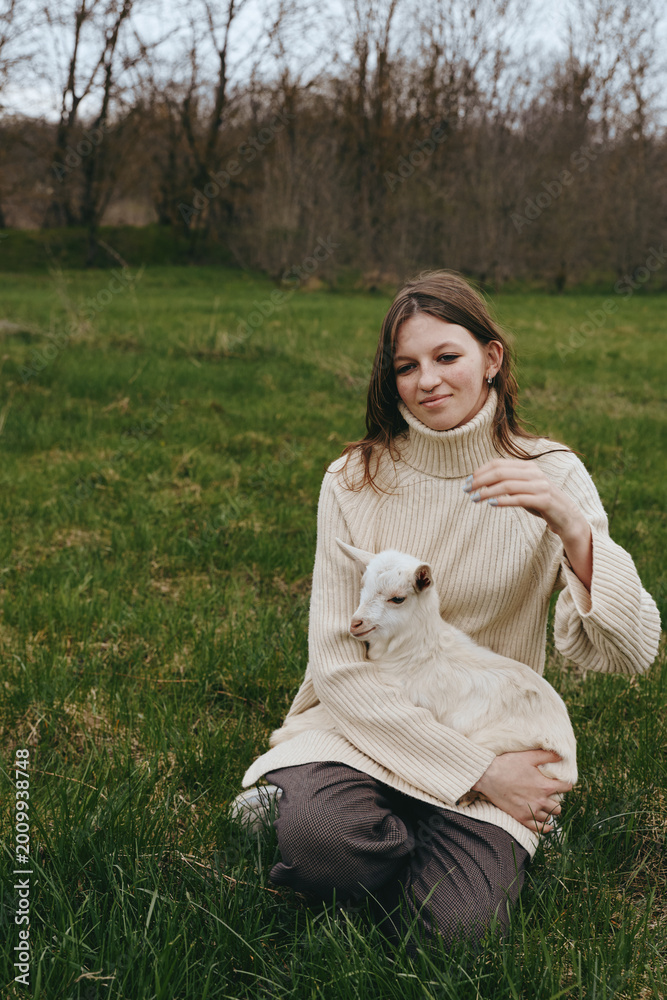 Obraz premium A woman in a cozy sweater sits in a grassy field, gently holding a young goat kid on her lap. A calm lifestyle moment capturing bond with animals and open outdoors.