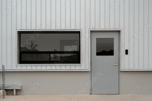 Slika na platnu Exterior of modern industrial building with white corrugated metal wall, closed grey door, and dark tinted window reflecting trees