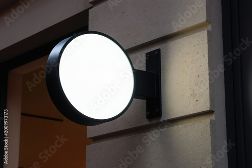 Round illuminated sign mockup hangs on modern building facade. Use for brand logo design. Modern sign outside shopfront. Exterior business sign.