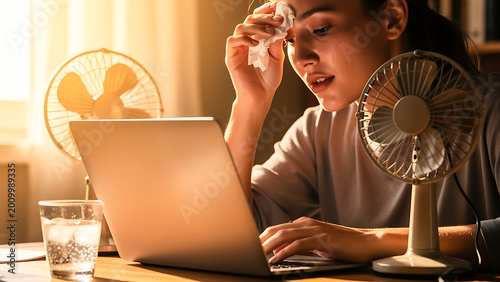 Woman wiping sweat while working on laptop in heat. Stressed worker feeling hot during summer day with electric fan. Dehydration and discomfort in office or home environment workplace.