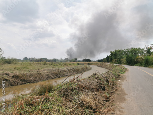 This image shows a rural area with a canal running beside a narrow road. In the distance, a fire is actively burning. Thick black smoke rises high into the sky from the flames. The smoke spreads acros