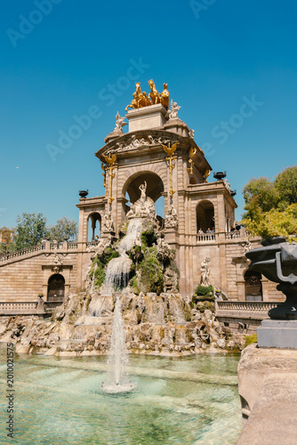Large stone monumental fountain with tiered waterfalls, classical sculptures, and a golden chariot statue at the top under a clear sky.
