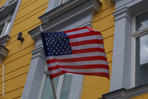 American flag waving in the wind against a yellow classic building facade. United States national symbol, patriotism, and diplomacy concept