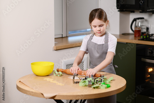Foto Little girl in apron rolling dough with wooden rolling pin, independent child co
