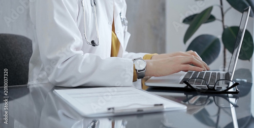 Obraz na plátně Doctor woman wearing white medical coat over a yellow blouse is working on laptop with stethoscope on the glass desk