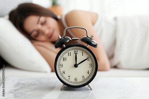 Young woman slepic peacefully in bed near alarm clock showing early morning time. Peaceful slumber, rest and waking up concept. Morning routine for healthy lifestyle.