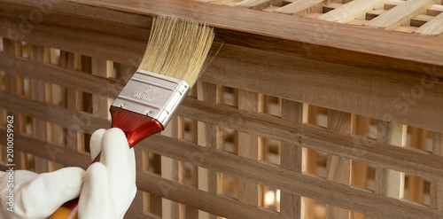 Paintbrush applies finish to an acacia wood lattice table. Gloved hand coats the slatted surface during furniture maintenance. DIY woodwork and home improvement concept..