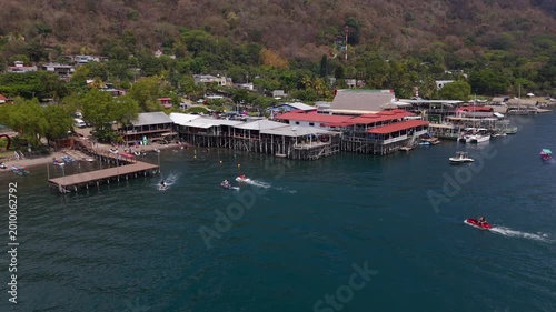 Aerial view of Lago de Coatepeque in El Salvador. Cinematic footage captures people jet skiing, paddleboarding, swimming, and relaxing on the water, with lakeside restaurants and hotels.