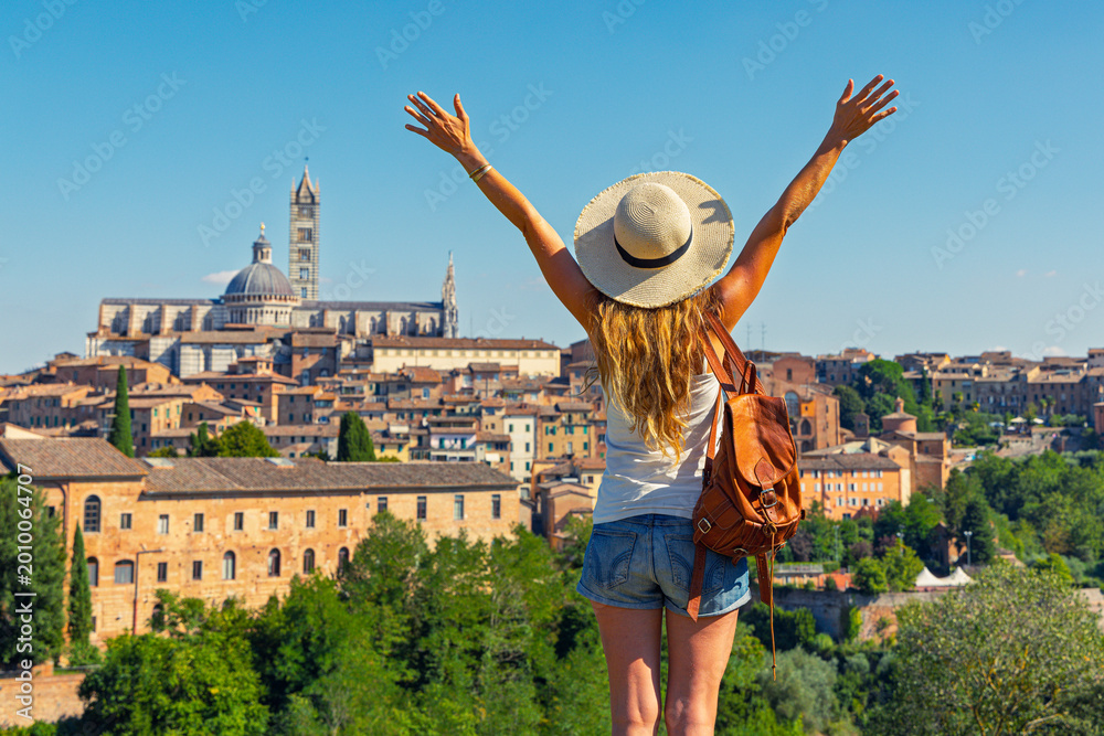 Fototapeta premium Happy young woman traveler with leather backpack and straw hat standing with arms raised and enjoying the scenic skyline view of siena, italy