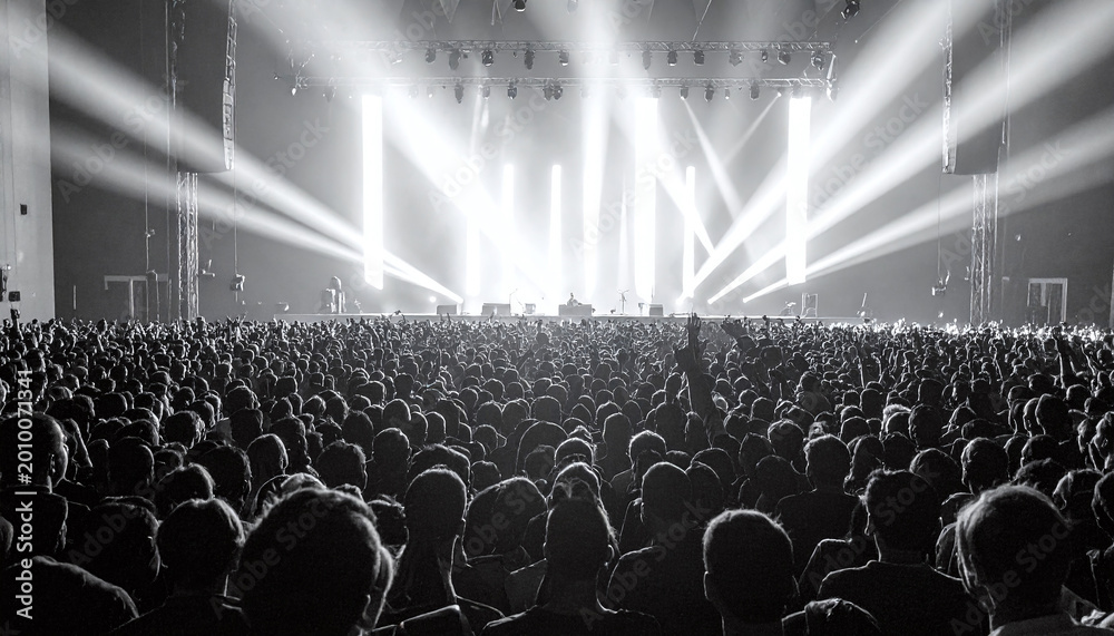Fototapeta premium Dramatic black and white shot of a massive crowd at a live music concert. Bright stage lights beam through the dark arena as fans cheer loudly. Ideal for event promotion and music themes.
