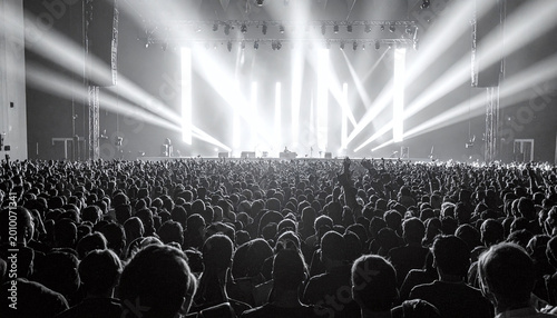 Dramatic black and white shot of a massive crowd at a live music concert. Bright stage lights beam through the dark arena as fans cheer loudly. Ideal for event promotion and music themes.