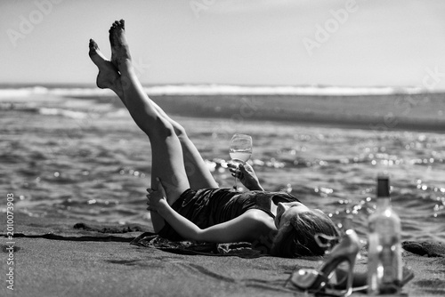 Konstfotografi Woman lying on sandy beach with legs raised holding wine glass
