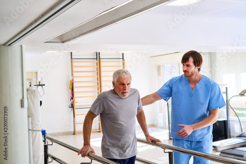 Quadro su tela Senior Man Walking With Physiotherapist Assistance On Parallel Bars In Rehab Cen