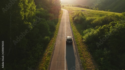 Car travels along a winding rural road. Sunlight bathes the scene in warm, golden hues. Lush green trees and grassy fields line both sides of the road. The vehicle moves steadily forward