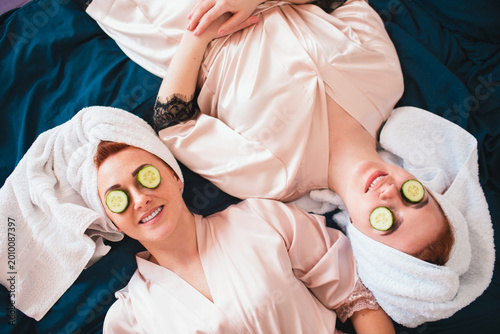 Two female models make an eye mask using fresh green cucumber. Two young women in towels and pajamas have a fun spa party together in home.