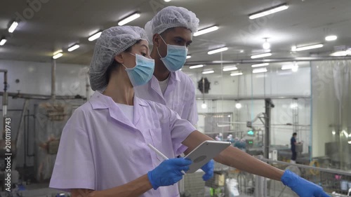 Two scientists in protective gear reviewing data on a tablet in a factory setting