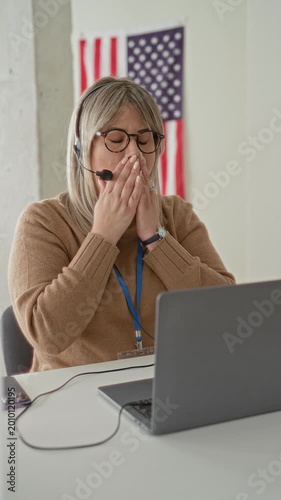 Woman yawning with hand over mouth at laptop in building wearing headset and voted sticker; civic duty tired.
