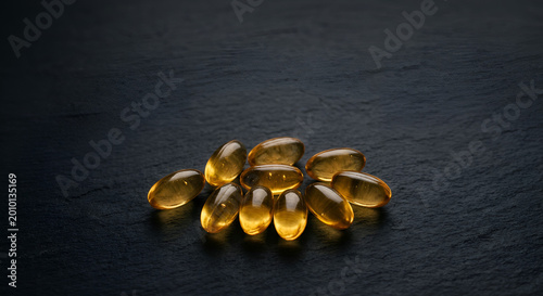 Supplements of golden yellow softgel capsules placed on a dark textured surface suitable for health and nutrition themes