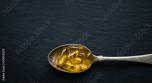 Close-up of a metallic spoon holding yellow fish oil capsules on a dark textured background emphasizing health supplements and