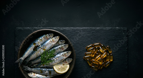 Fresh sardines garnished with lemon and herbs alongside omega-3 capsules on a dark slate background