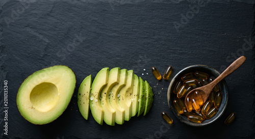Freshly sliced avocado with a bowl of omega-3 fish oil supplements on a dark slate background