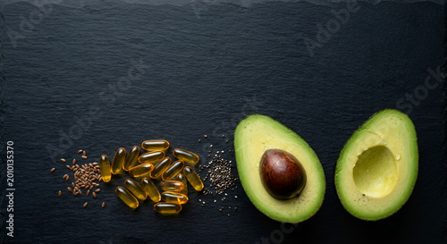 fresh ripe avocado with seed and smooth-textured capsules of fish oil supplement pills on dark slate background