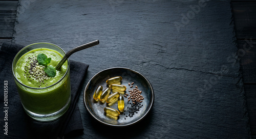 Healthy green smoothie garnished with mint alongside supplement capsules and seeds on a dark textured surface