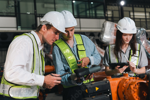 Robot engineer team auto factory inspection with tablet and clipboard, focused worker collaboration under safety helmet and vest around modern machine arm indoors industrial technology