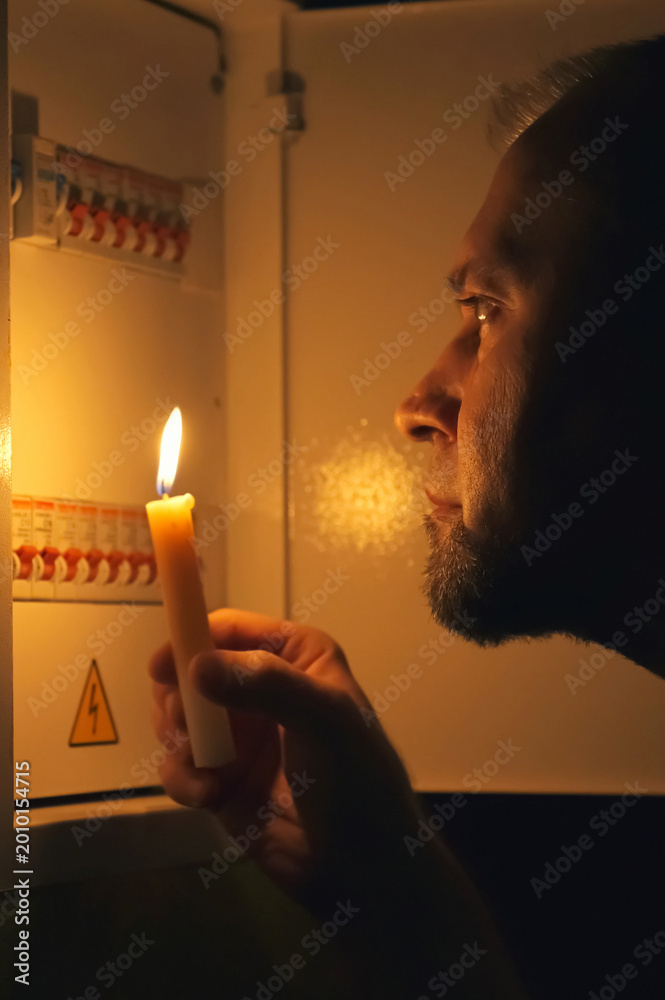 Fototapeta premium Man with Candle Inspecting Electrical Switchboard During Power Outage at Home